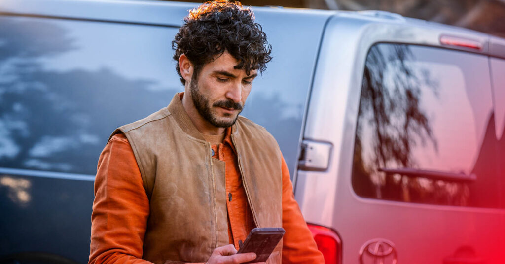 En man tittar ner i sin telefon. I Bakgrunden står en bil.