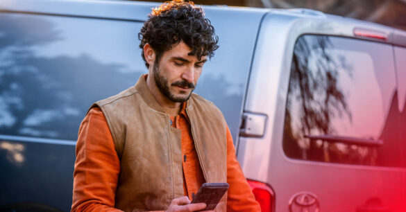 En man tittar ner i sin telefon. I Bakgrunden står en bil.